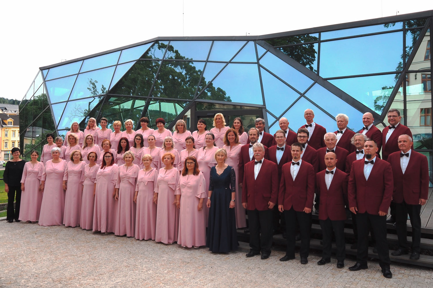 Gruppenfoto eines Chores, links die Damen in langen altrosafarbenen Kleidern, rechts die Herren in weinroten Sakkos, im Hintergrund ein modernes Gebäude aus Glas