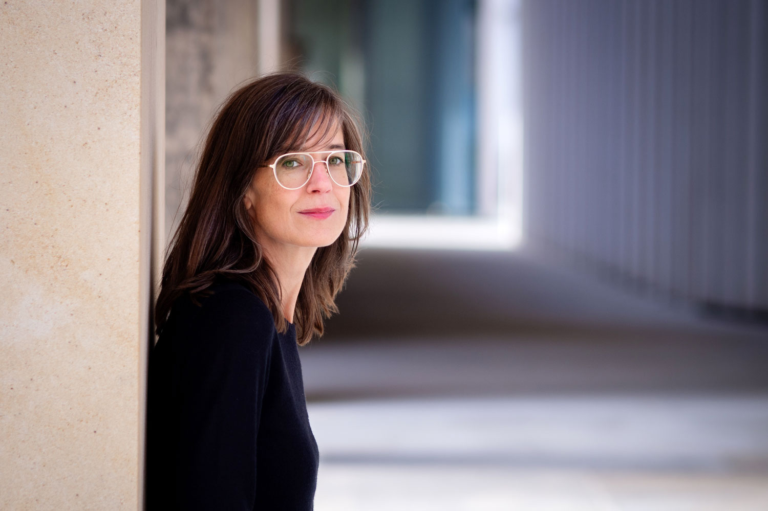 Prof. Andrea Tober lehnt in einem längeren Gang links an einer Wand und schaut in die Kamera. 