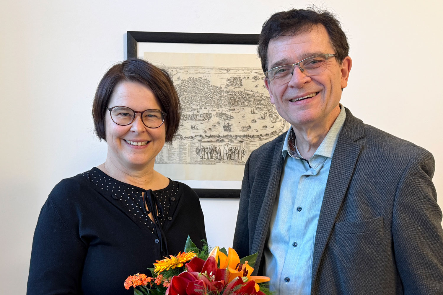 Eine Dame und ein Herr blicken lächelnd in Richtung Betrachter. Im Vordergrund sieht man einen Blumenstrauß, der gerade übergeben wurde.